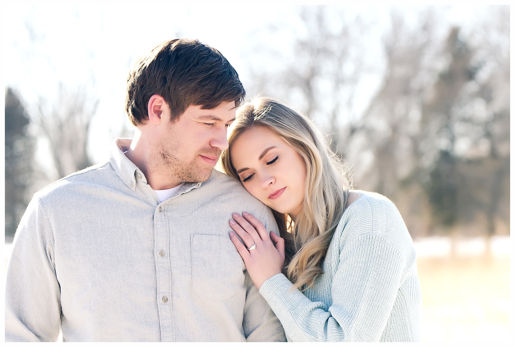 couple wearing light blues and girl rests head on fiancé's shoulder in the snow.  