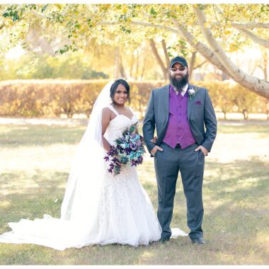 Bride and groom in purple and grey standing together under fall trees