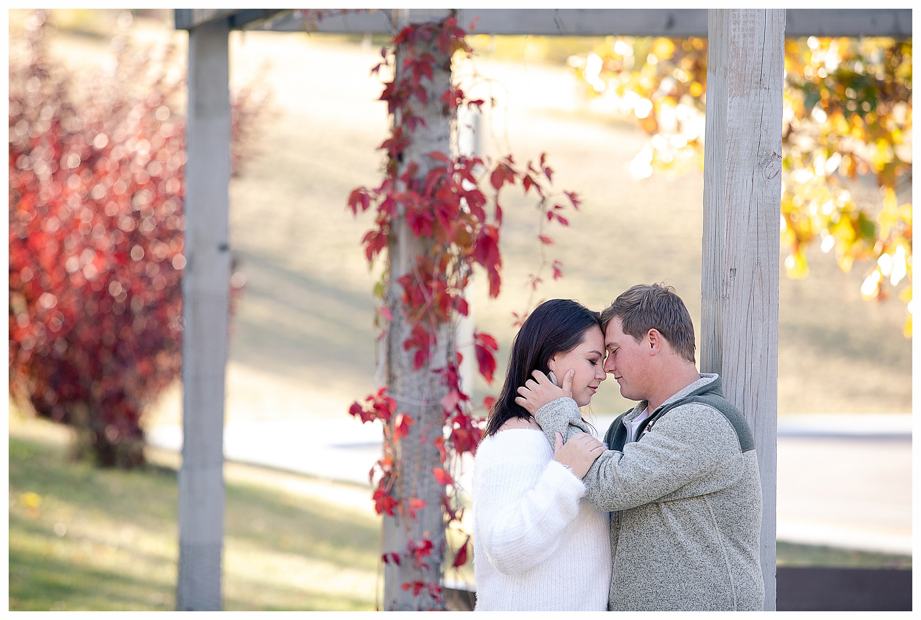 couple stand with foreheads touching under arbor with red and gold fall trees