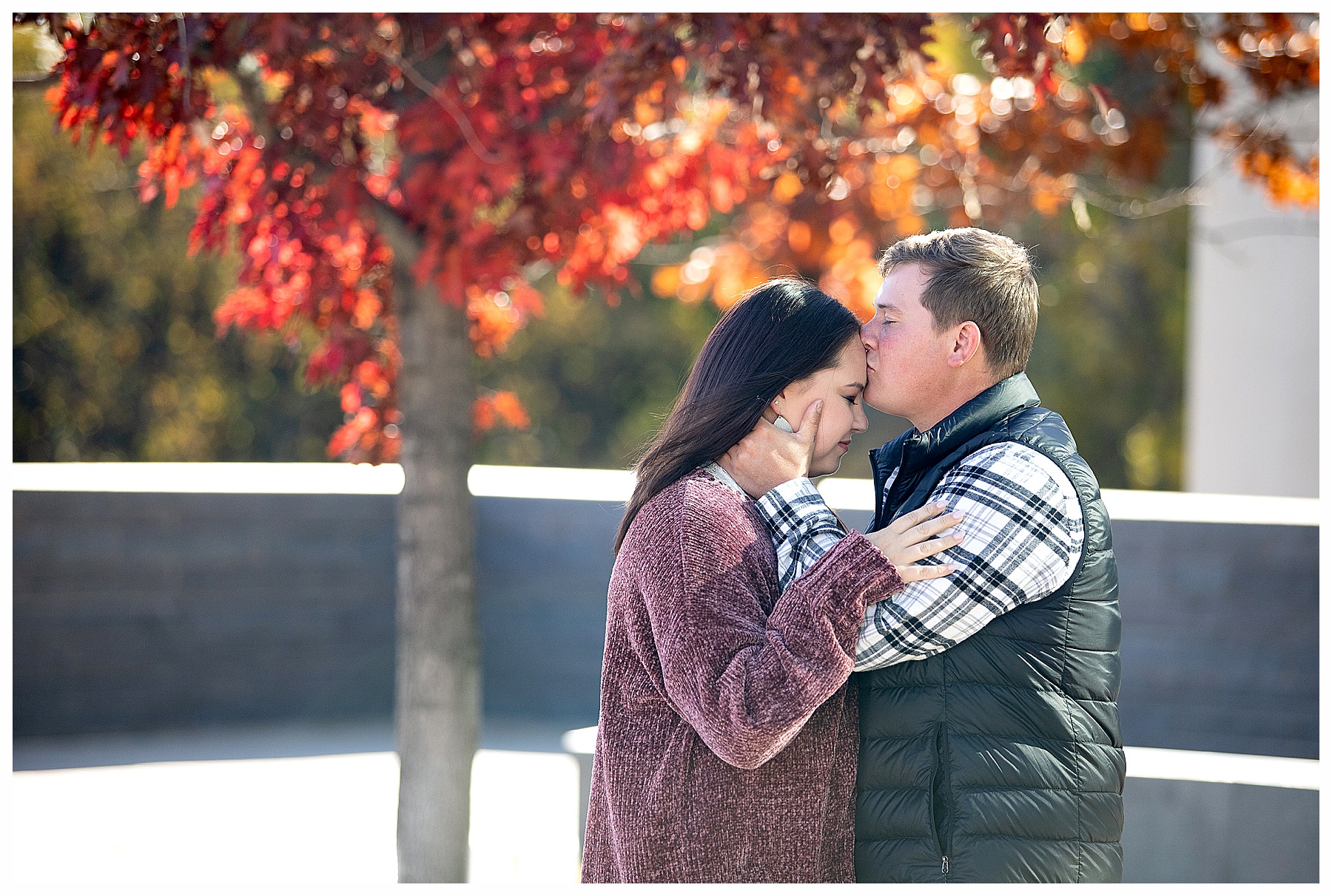 Man kisses fiancé on forhead under red fall trees at Bismarck Heritage Center