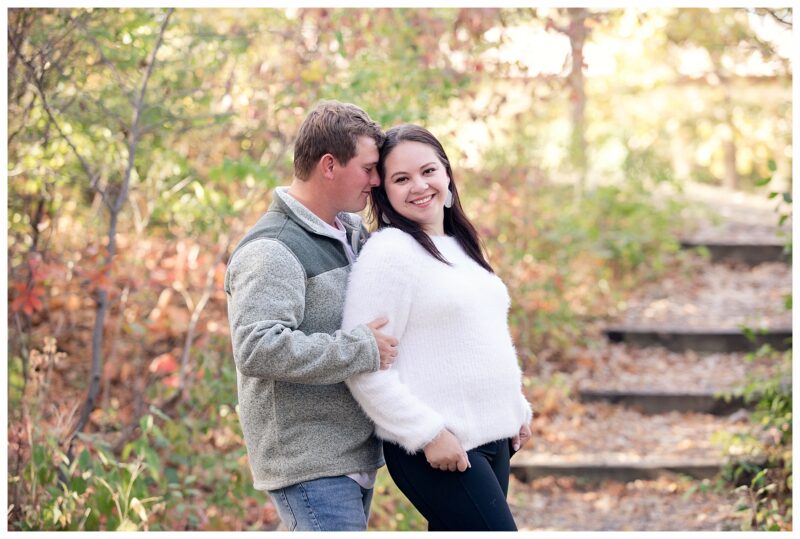 Engaged couple stand on wood steps under fall trees at Sleepy Hollow Park in Bismarck ND