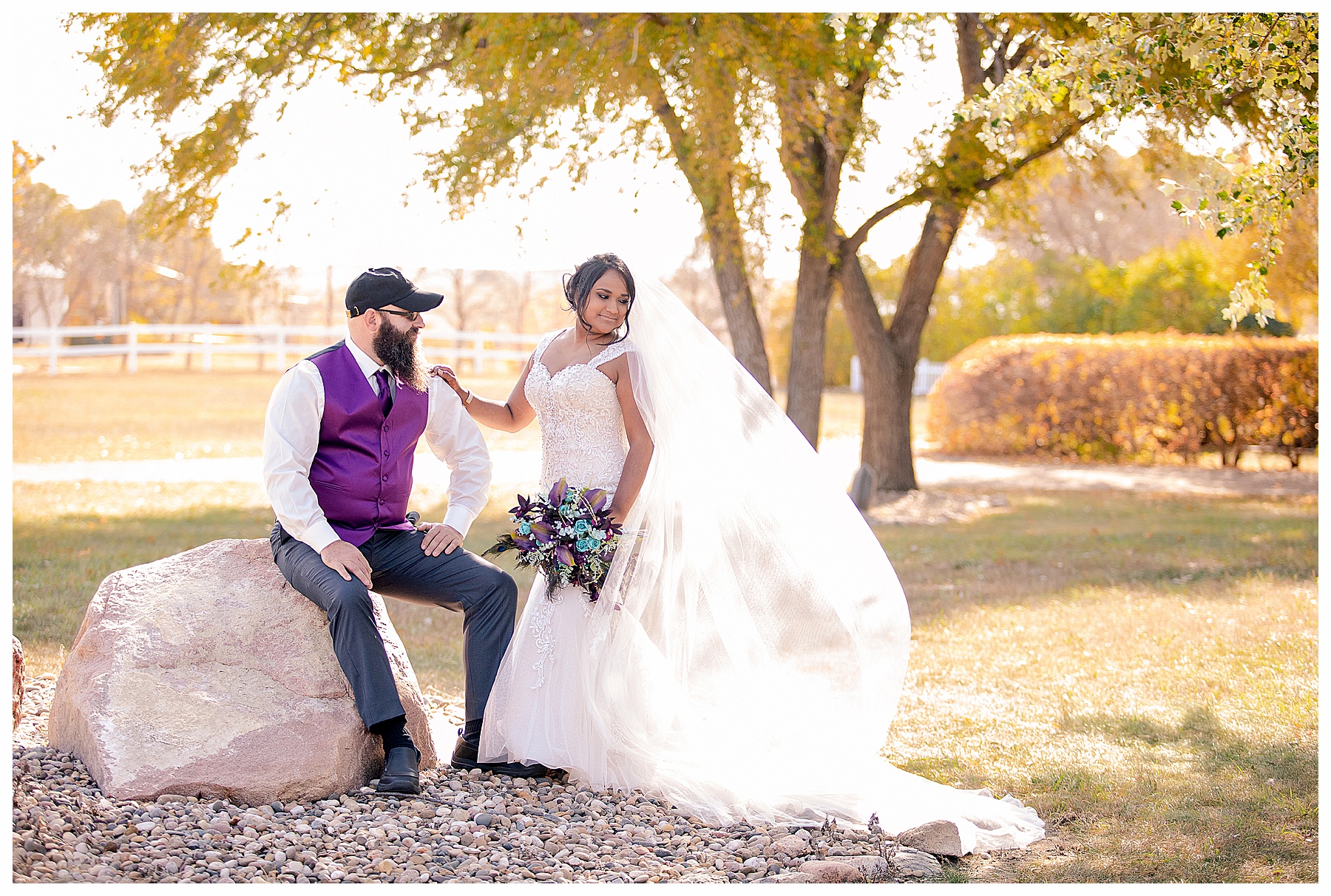 Groom sits on boulder as he looks at his bride with blowing veil