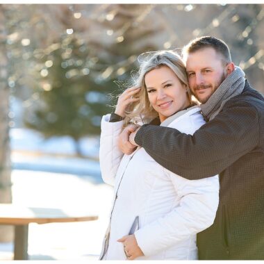 Engaged couple take photo with snow and white lights in background. Bride wearing white jacket