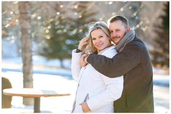 Engaged couple take photo with snow and white lights in background. Bride wearing white jacket